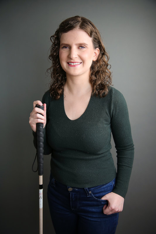Photo of Sammi Grant from knees up. She is a white woman in the early 30s with brown, curly hair and grey-eyes holding a white cane.